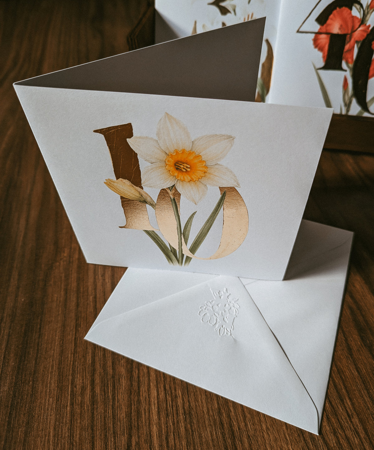 A white greeting card standing open on a wooden surface, printed with a daffodil and the number '10'' in stylised lettering. A matching white envelope lies in front, and part of a branded box is visible in the background.