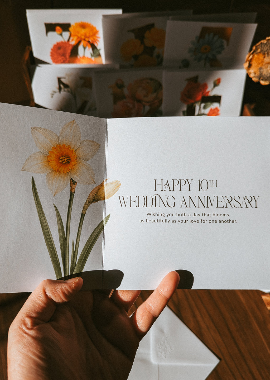 A hand holds an open white greeting card with a watercolor daffodil illustration and the text 'HAPPY 10TH WEDDING ANNIVERSARY.' Other floral greeting cards are stacked in the background on a wooden surface.