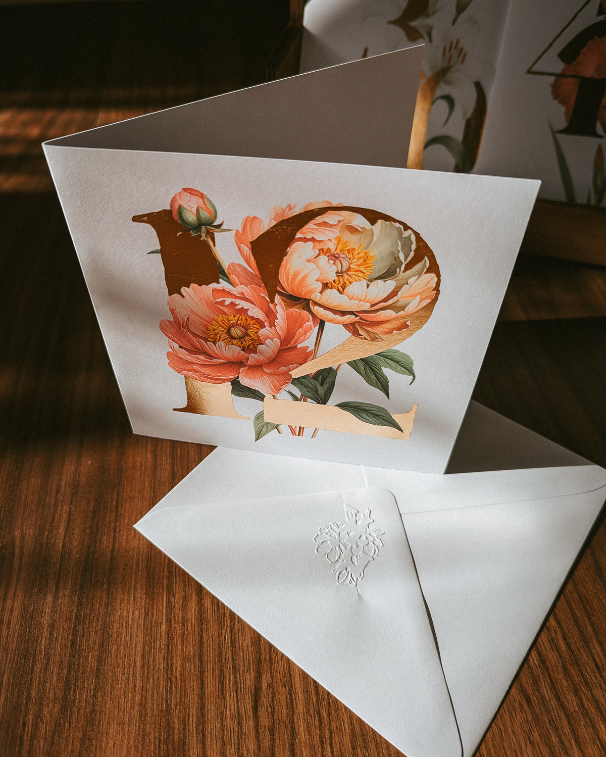 A square greeting card with a vintage-style floral illustration of the number 12 in peach, orange, and green tones, displayed on a wooden surface with a white embossed envelope in front.