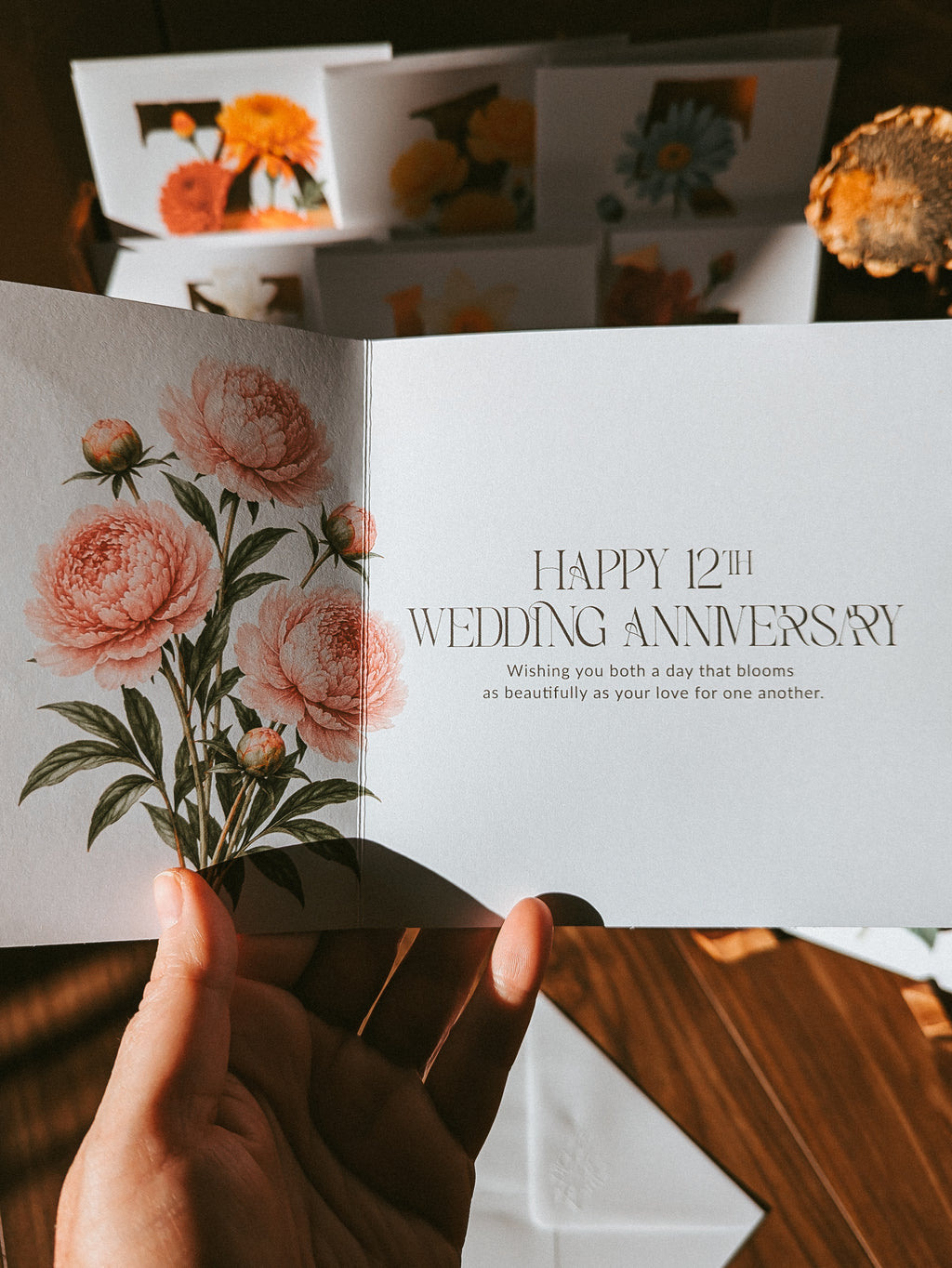 An open floral anniversary card held in a hand, showing the message 'HAPPY 12TH WEDDING ANNIVERSARY' with pink peony illustrations. Other matching cards are stacked in the background on a wooden surface.
