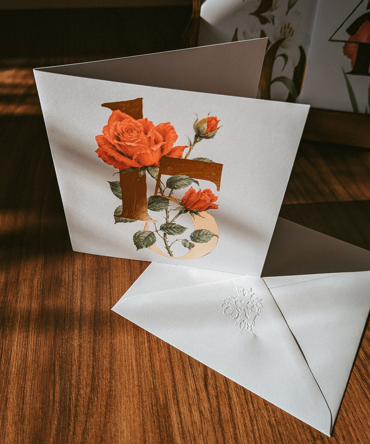 A square white greeting card with a large decorative gold number '15' overlaid with red roses and green leaves, shown open on a wooden surface with a matching white envelope in front.