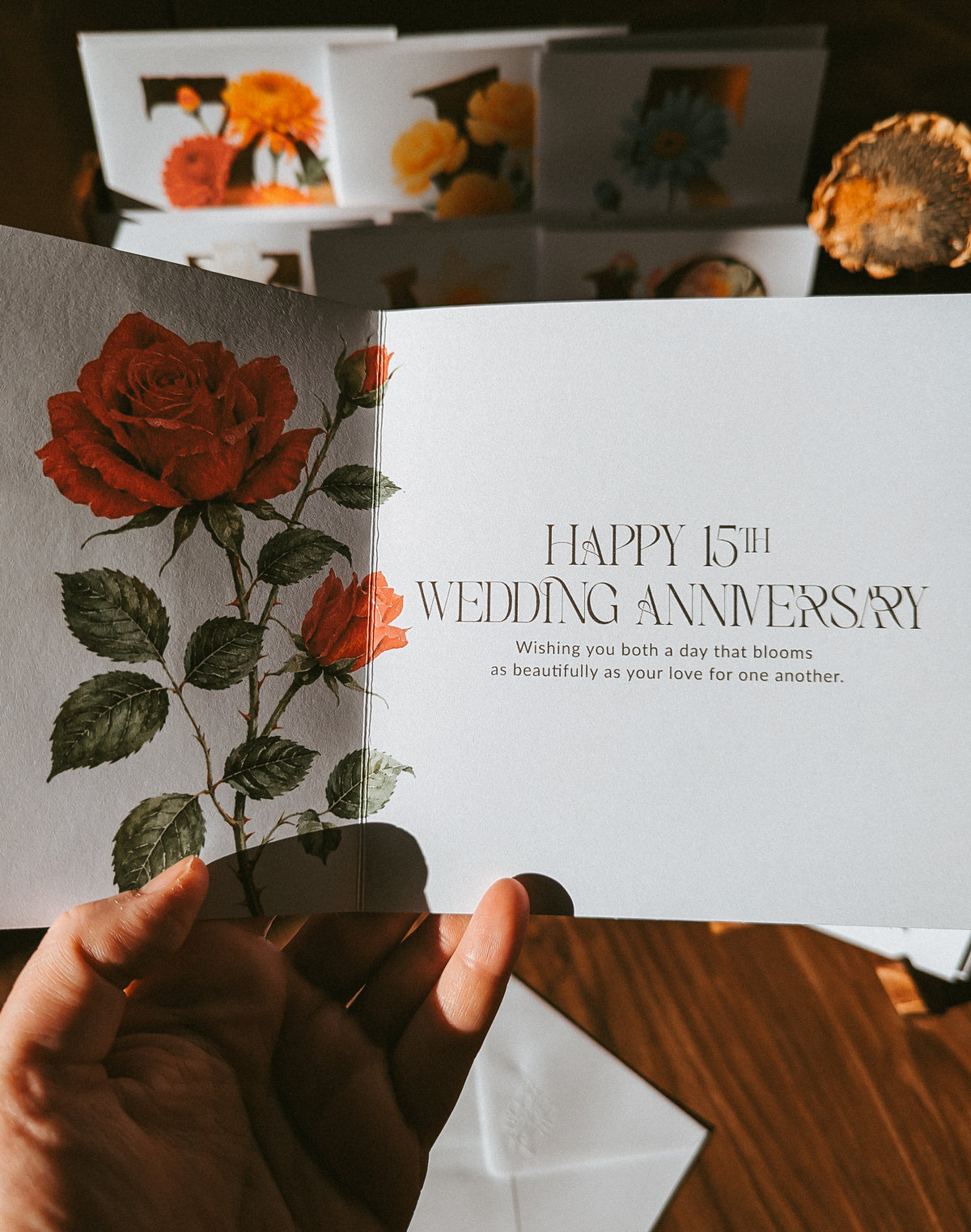 A hand holds an open greeting card with a white interior featuring the text 'HAPPY 15TH WEDDING ANNIVERSARY' and a large red rose illustration. Other floral greeting cards are stacked in the background on a wooden surface.
