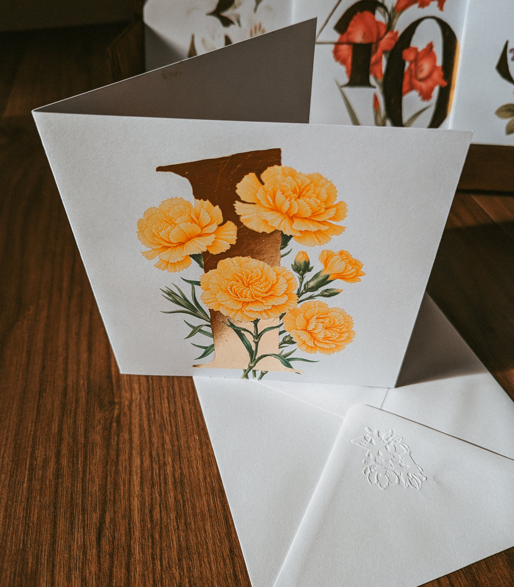 A white greeting card with a large gold '1' and yellow-orange carnation flowers, shown standing open on a wooden table with white envelopes in front.