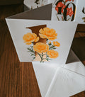 A white greeting card with a large gold '1' and yellow-orange carnation flowers, shown standing open on a wooden table with white envelopes in front.
