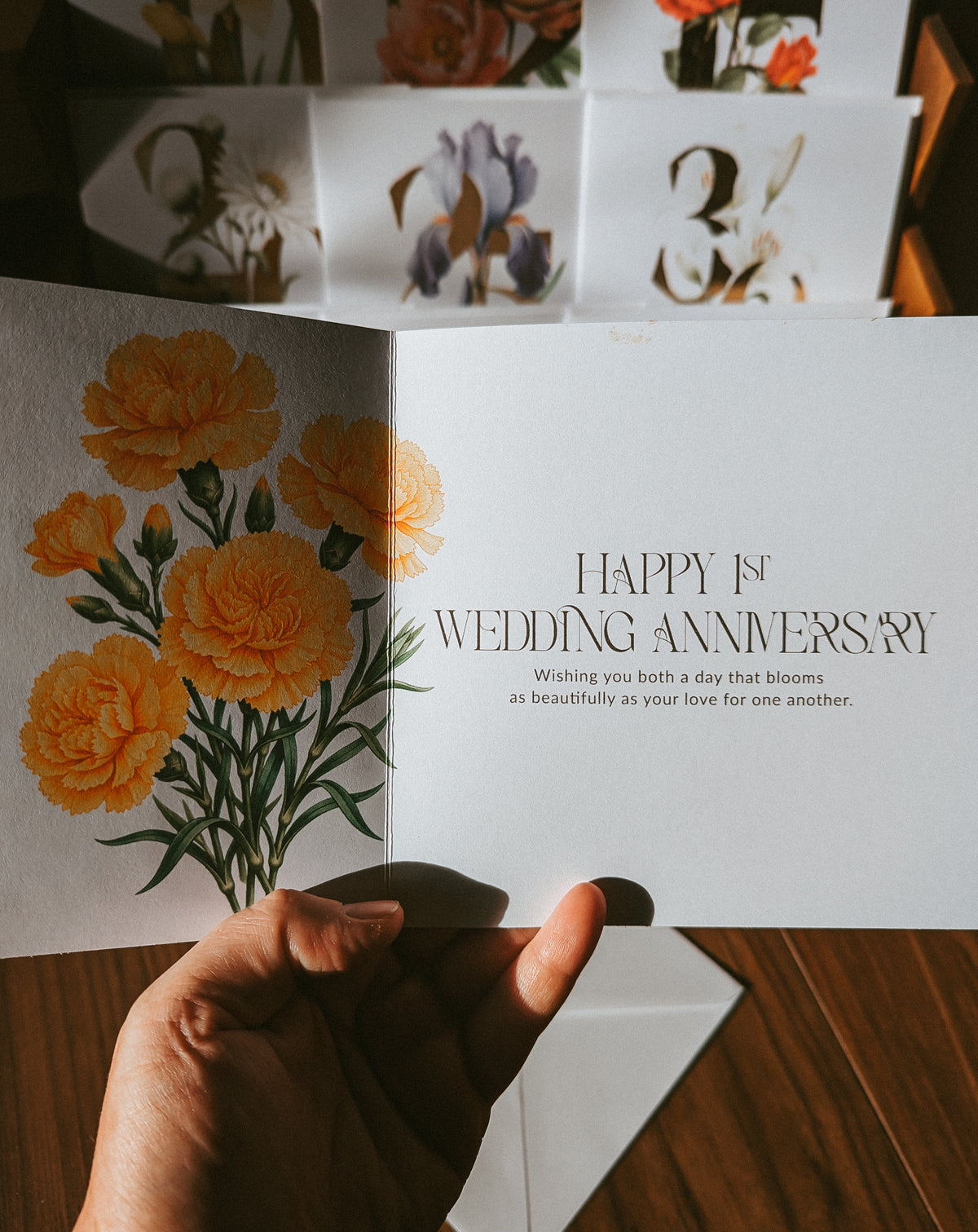 An open greeting card held in a hand, showing a watercolor-style yellow flower illustration on the left page and the text 'HAPPY 15th WEDDING ANNIVERSARY' on the right page. More floral greeting cards are stacked in the background on a wooden surface.
