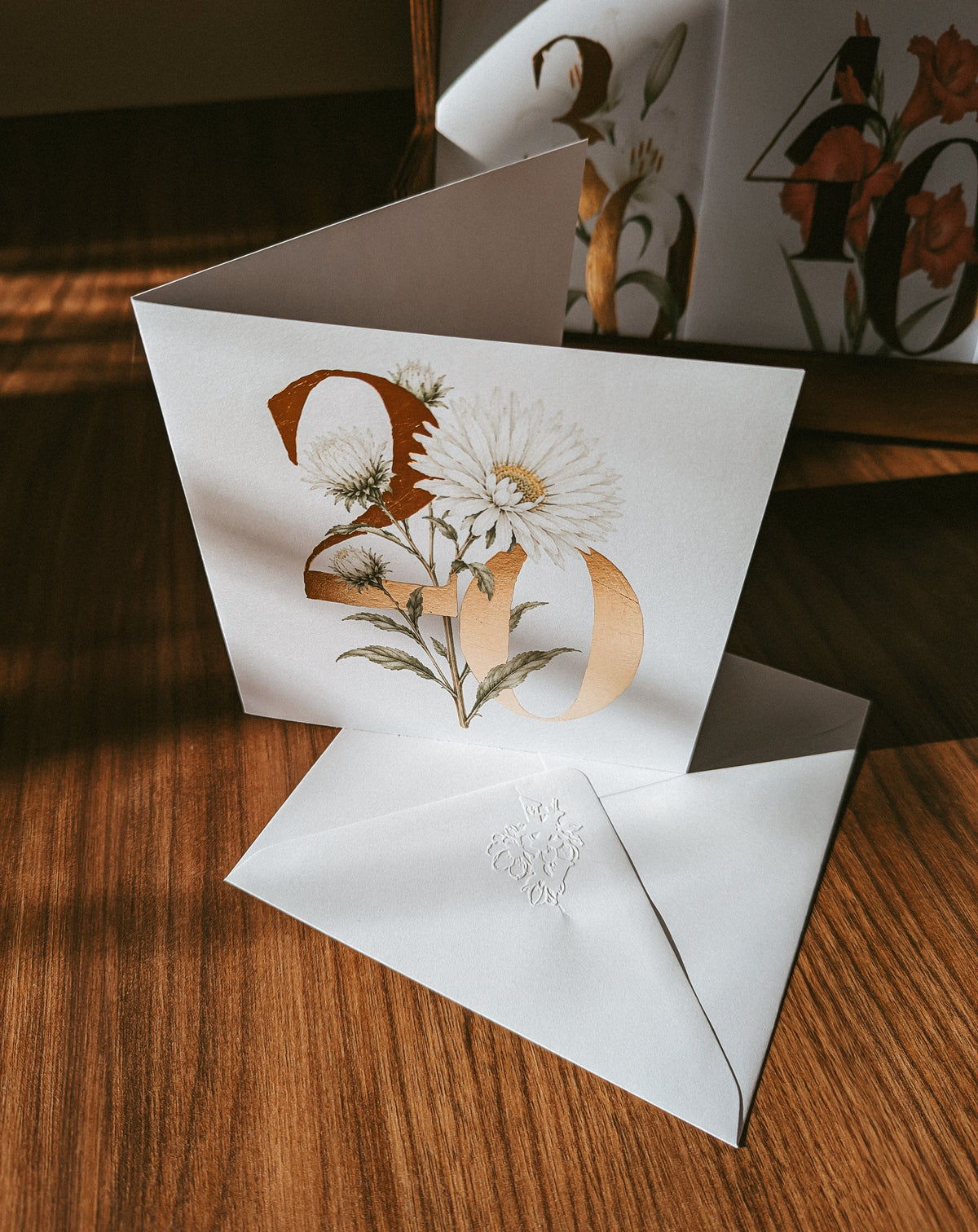 A white wedding invitation card with a botanical-style gold and white floral design over a large gold number '20', shown standing on a wooden table with a matching white envelope in front.
