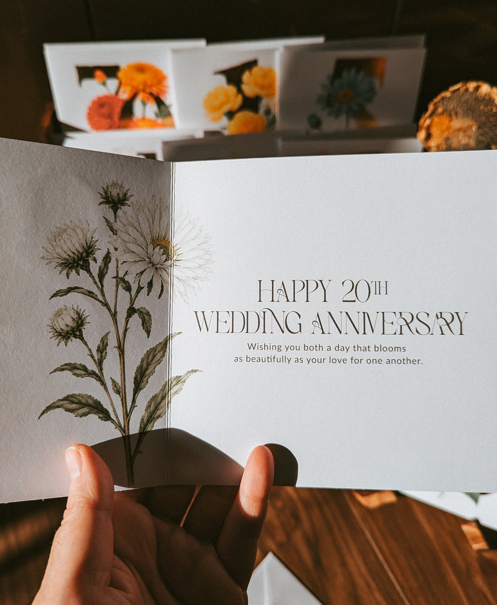 A hand holds an open wedding anniversary card with a botanical illustration on the left page and elegant text on the right page reading 'HAPPY 20TH WEDDING ANNIVERSARY' and a short message below. The background shows more floral greeting cards and a warm wooden surface.
