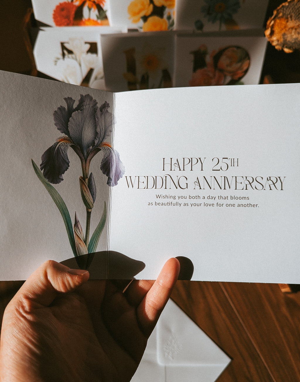 A hand holds an open greeting card showing a watercolor-style iris illustration on the left panel and the text 'HAPPY 25th WEDDING ANNIVERSARY' on the right panel. Other floral greeting cards are stacked in the background on a wooden surface.