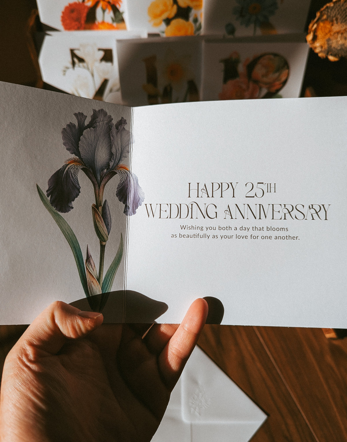 A hand holds an open greeting card showing a watercolor-style iris illustration on the left panel and the text 'HAPPY 25th WEDDING ANNIVERSARY' on the right panel. Other floral greeting cards are stacked in the background on a wooden surface.