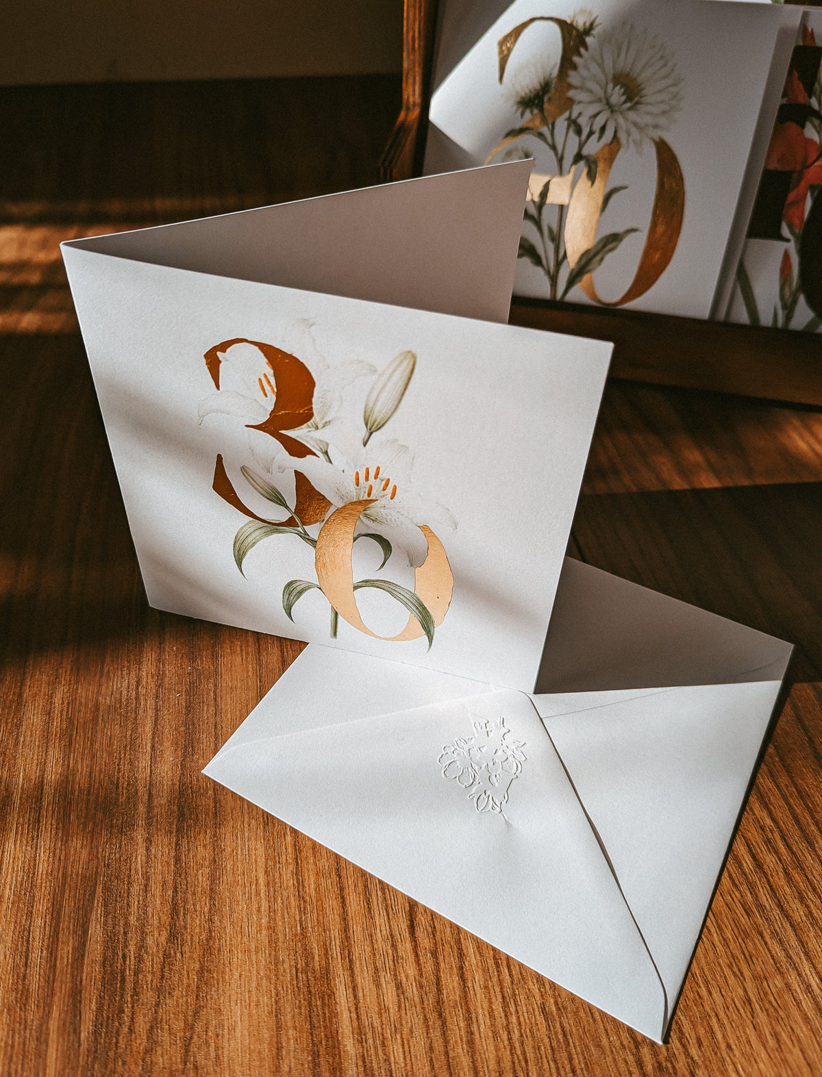 A white greeting card with a large metallic gold floral monogram and lily illustration, shown open on a wooden table next to a matching white envelope with an embossed monogram.