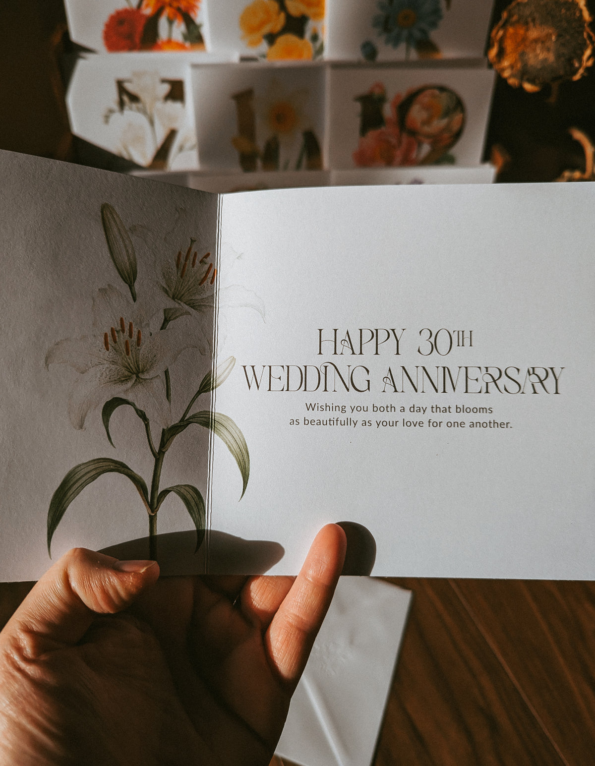 A hand holds an open greeting card with white lilies on the left page and the text 'HAPPY 30TH WEDDING ANNIVERSARY' on the right panel. More floral greeting cards are stacked in the background on a wooden surface.
