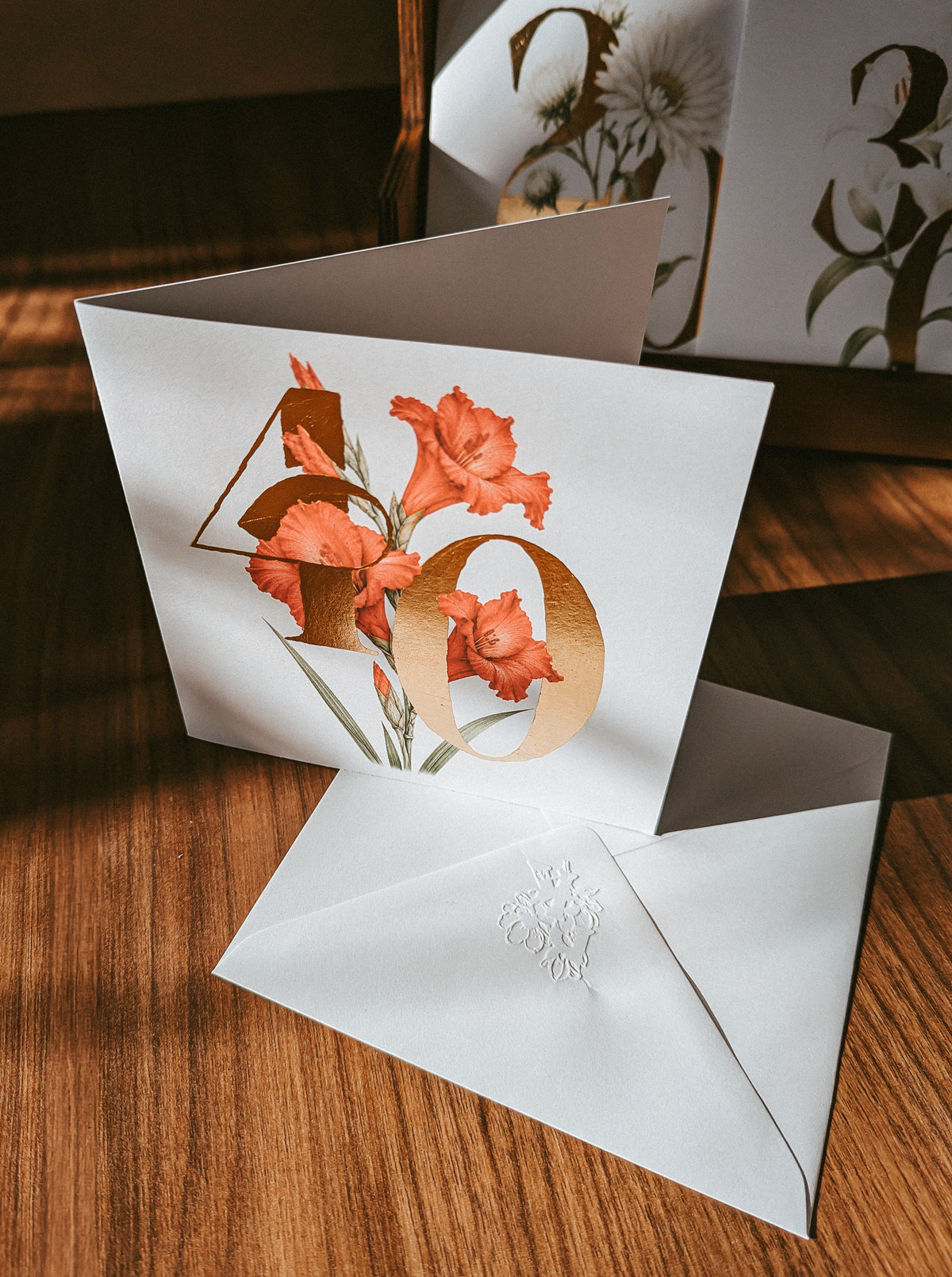 A white greeting card with a large metallic gold '40' and red floral illustration, shown open on a wooden surface with a matching white envelope in front.