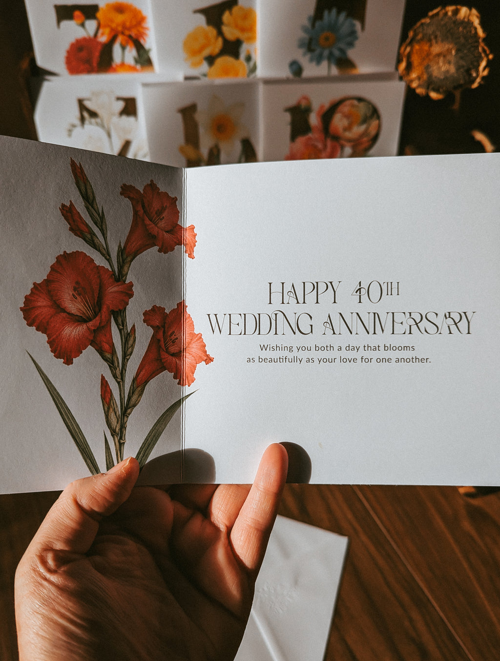 A hand holds an open greeting card with a floral illustration on the left page and the text 'HAPPY 40TH WEDDING ANNIVERSARY' on the right page. Other cards are stacked in the background on a wooden surface.