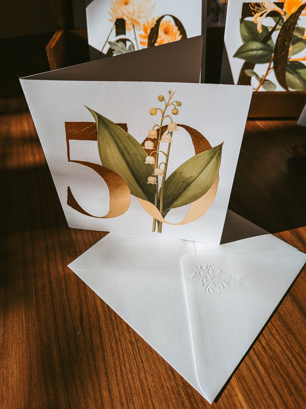 A white card displaying '50' with a lily of the valley floral illustration, shown standing upright on a wooden table with a matching white envelope in front.