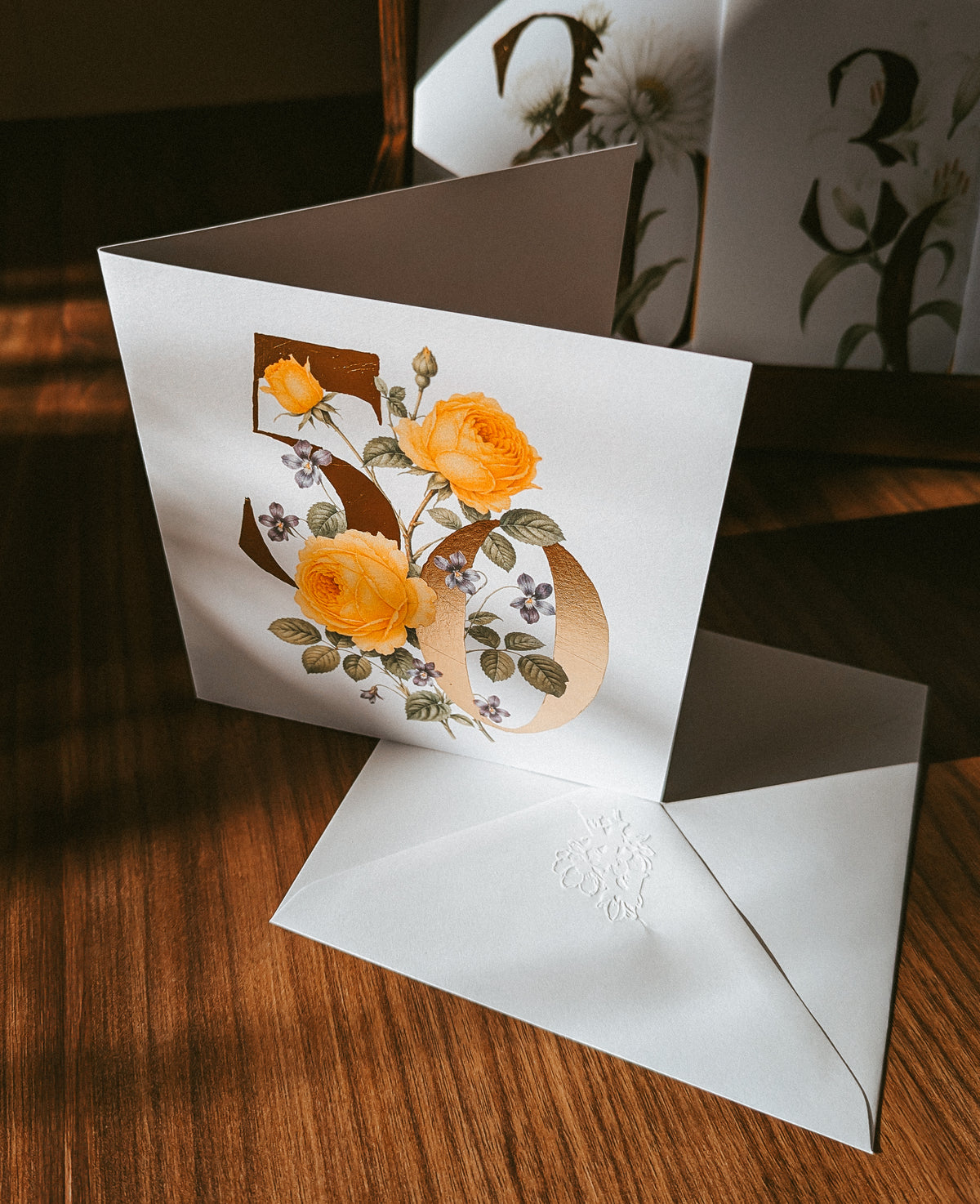 A white greeting card with a large gold number 50 decorated with yellow roses and green leaves, shown standing on a wooden surface with a matching white envelope in front.