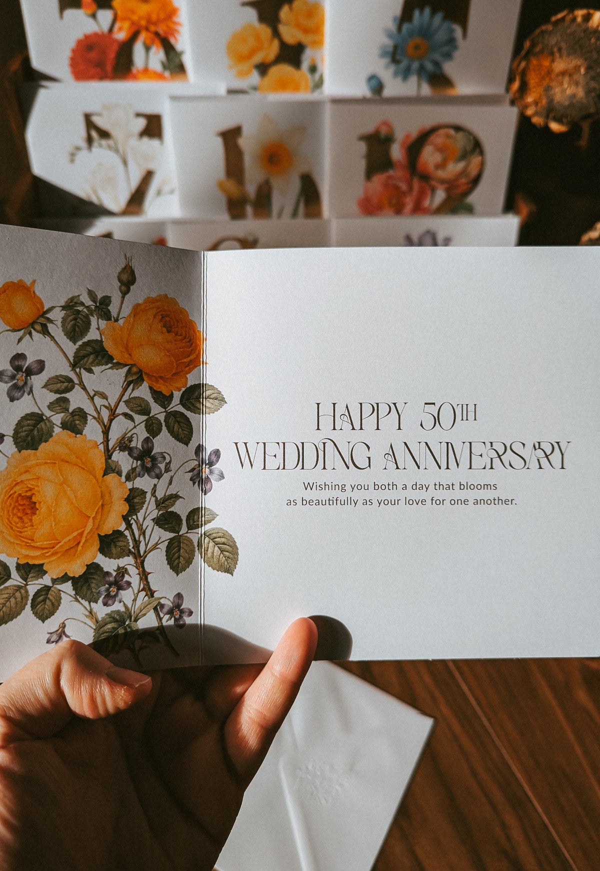 A hand holds an open greeting card with a floral illustration on one page and the text 'HAPPY 50TH WEDDING ANNIVERSARY' on the other. More floral greeting cards are stacked in the background on a wooden surface.