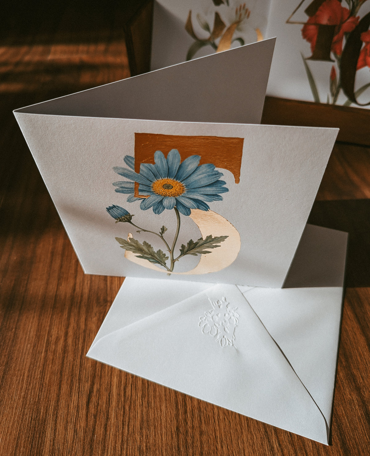 A folded greeting card with a blue flower illustration and a white embossed envelope, displayed on a wooden surface.