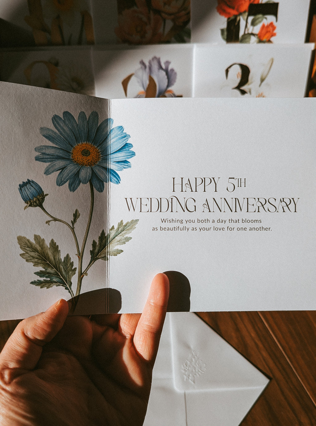 A hand holds an open wedding anniversary card featuring a blue aster illustration and the text 'HAPPY 5th WEDDING ANNIVERSARY' on a white card, with a matching white envelope on a wooden surface.