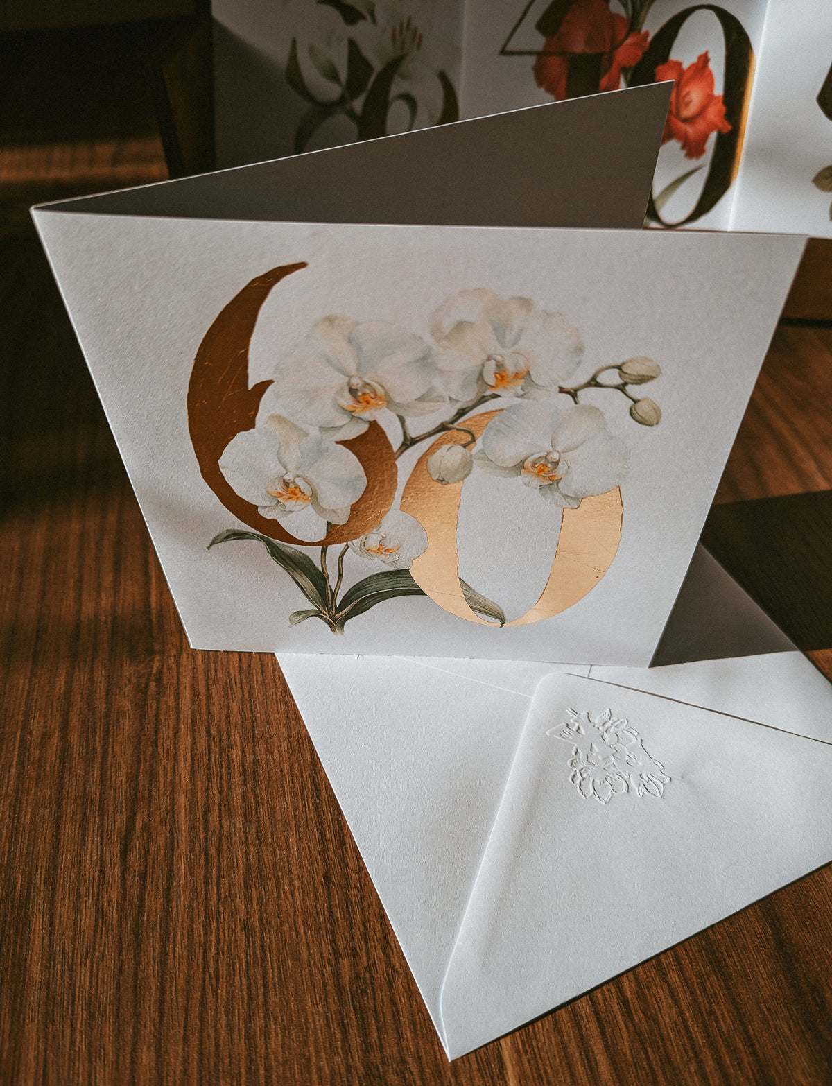 A white greeting card with a large metallic gold '60' and white orchid flowers, displayed on a dark wood table next to a matching white embossed envelope.