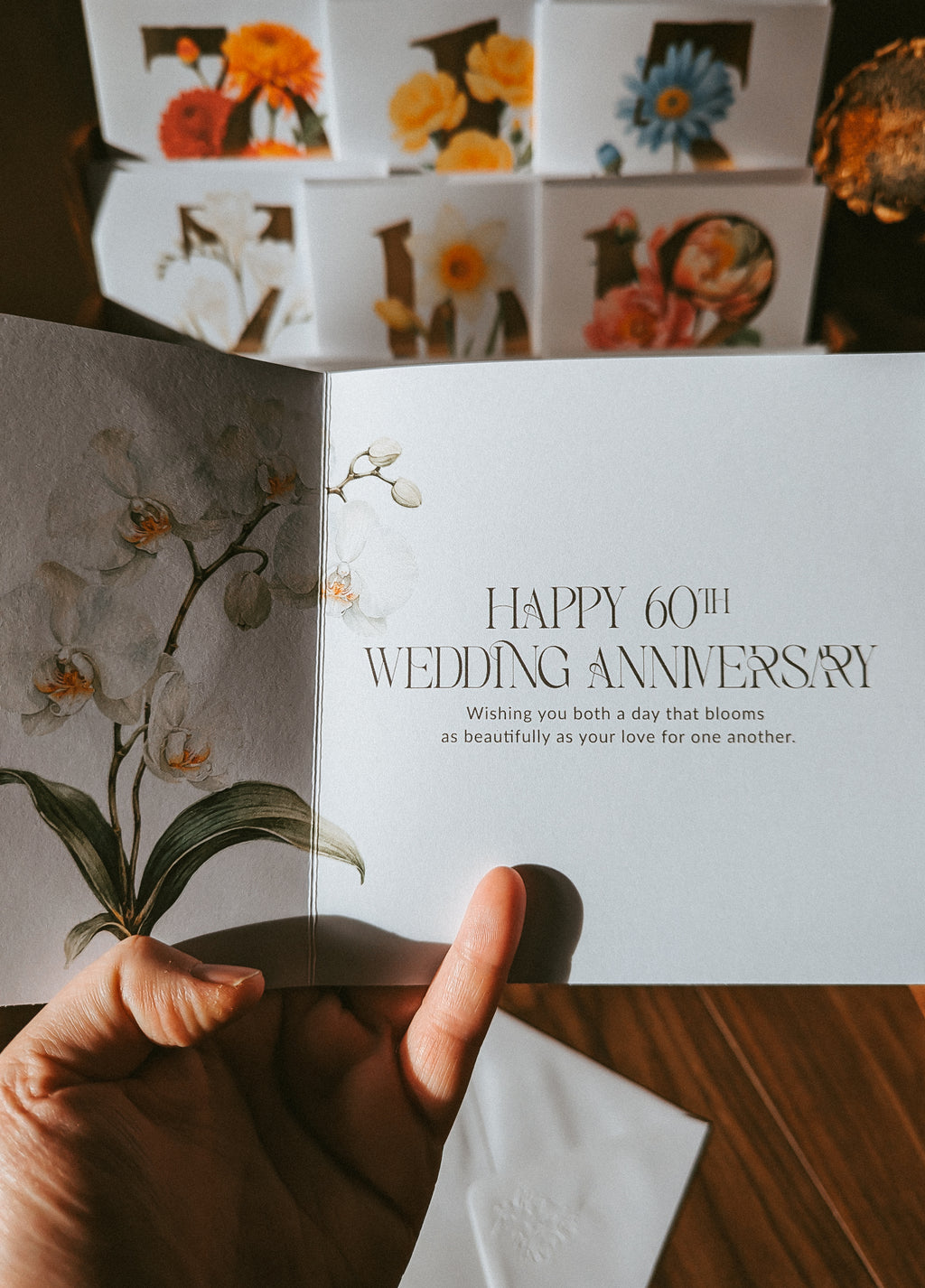 A hand holds an open greeting card with floral artwork and the text 'HAPPY 60TH WEDDING ANNIVERSARY' on a white page, with a matching stack of floral cards blurred in the background on a wooden surface.