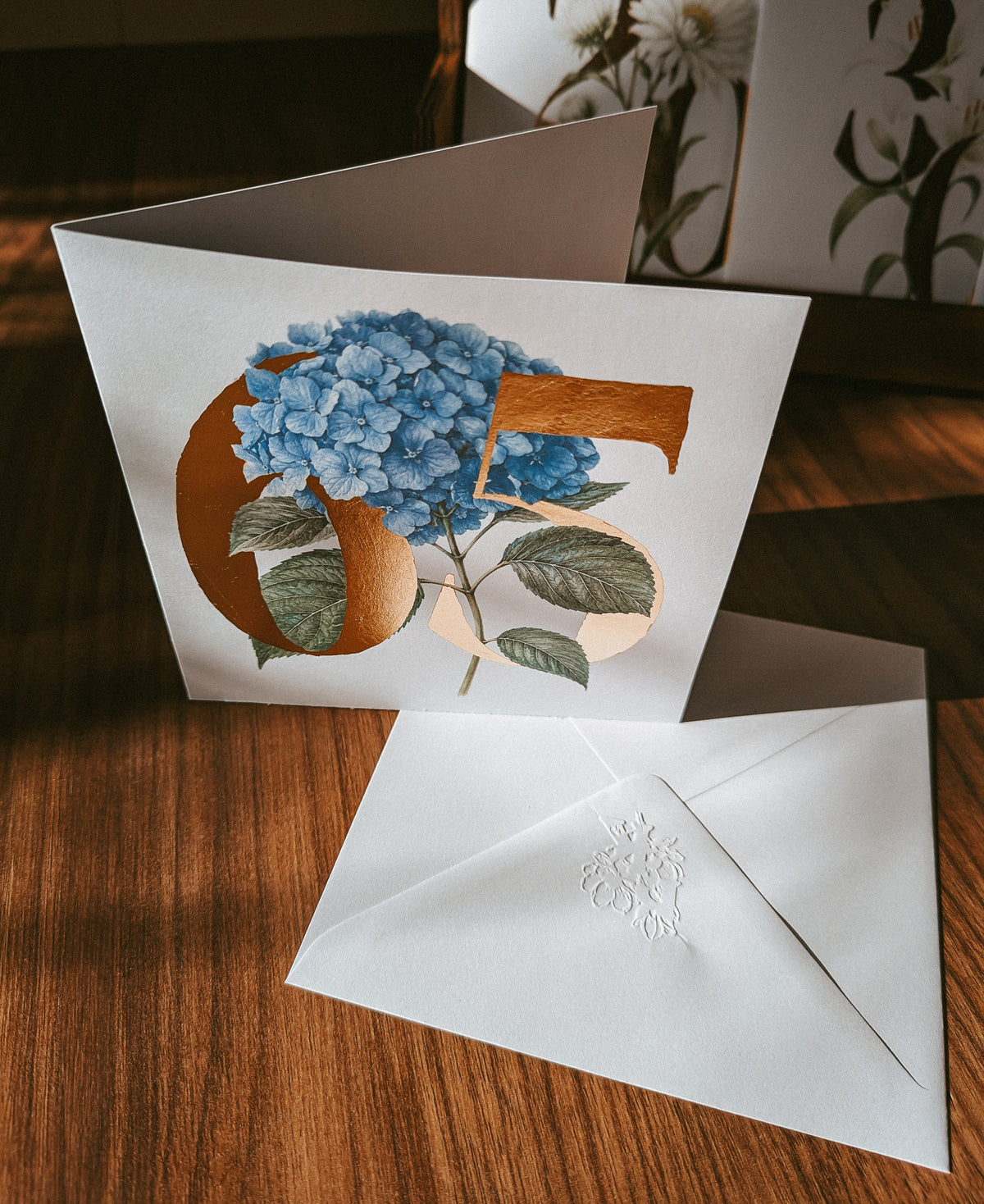 A white greeting card with a large gold number 65 and blue hydrangea flowers, shown open on a wooden table with a matching white embossed envelope in front.