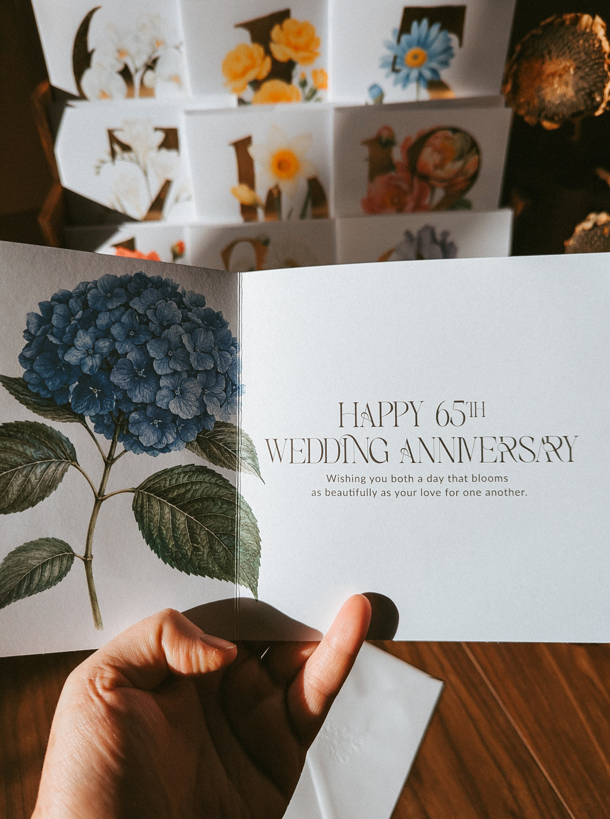 A hand holds an open wedding anniversary card featuring a blue hydrangea illustration and elegant serif text reading 'HAPPY 65th WEDDING ANNIVERSARY.' The background shows a stack of floral greeting cards on a wooden surface.