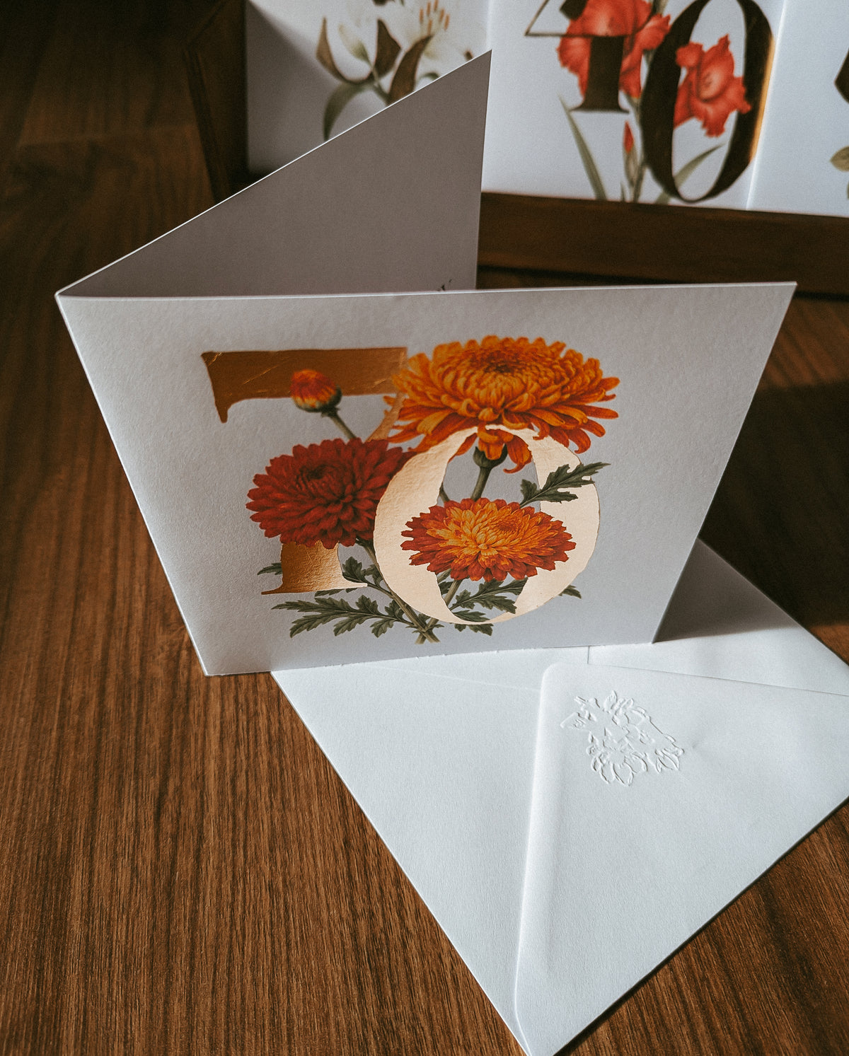 A folded greeting card with a large floral '70' design in orange, red, and cream tones, displayed open on a wooden surface with a white envelope in front.