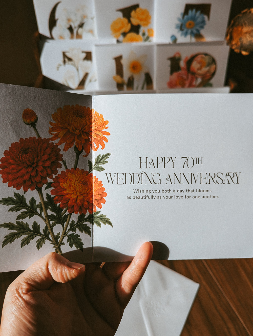A hand holds an open greeting card with orange flowers on the left panel and the text 'HAPPY 70TH WEDDING ANNIVERSARY' on the right panel. More floral greeting cards are stacked in the background on a wooden surface.