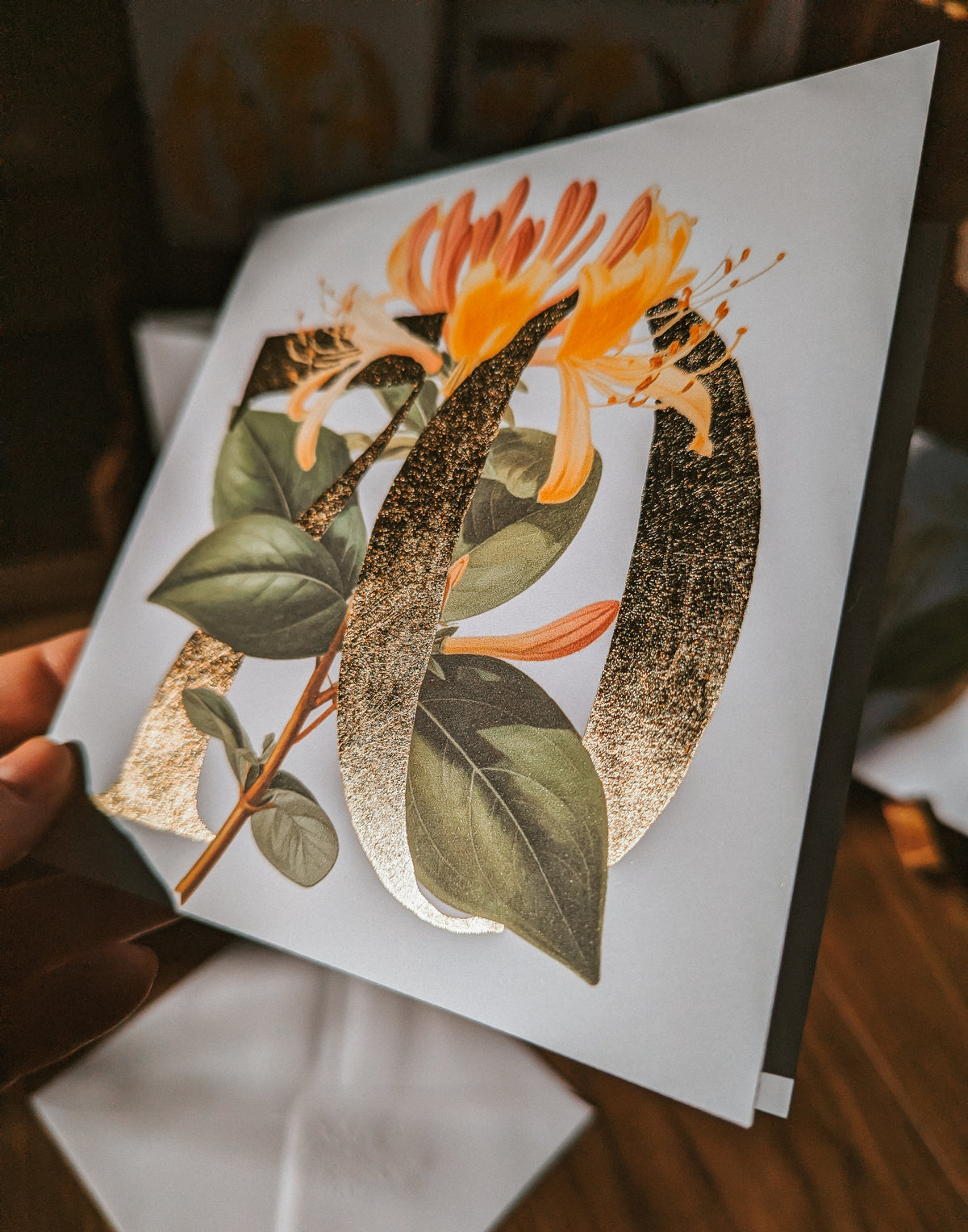 A close-up of a hand holding a square white card or print featuring a botanical illustration with orange flowers, green leaves, and a large metallic gold monogram letter '70' on a dark wood surface.
