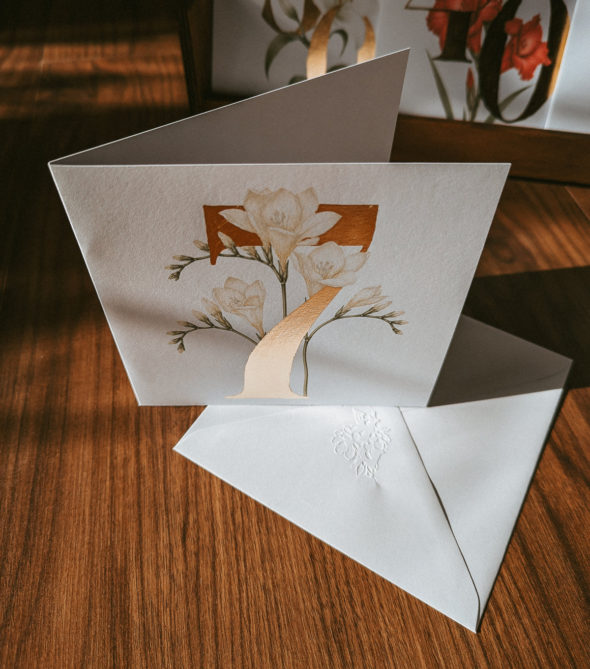 A folded white greeting card with a floral illustration and a large gold numeral '7' on the front, shown open on a wooden surface with a matching white embossed envelope in front.