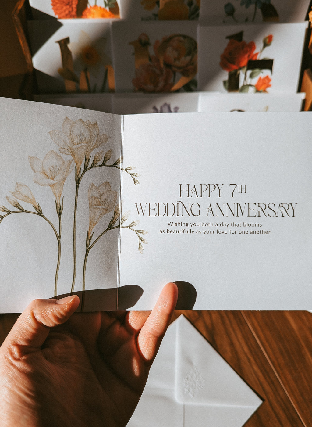 A hand holds an open white anniversary card with delicate floral illustrations and the text 'HAPPY 7TH WEDDING ANNIVERSARY' on a wooden surface, with more floral cards blurred in the background.
