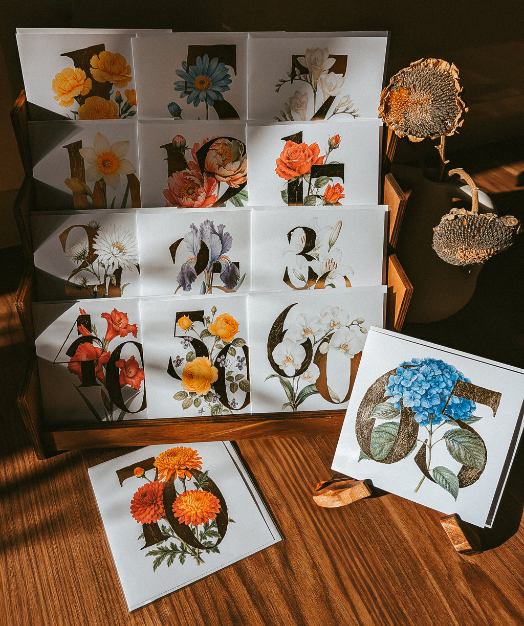 A set of floral and botanical greeting cards arranged on a wooden surface, with a folded card display showing multiple illustrated designs and two cards placed in front. The cards feature flowers and large numbers in artistic botanical illustrations.