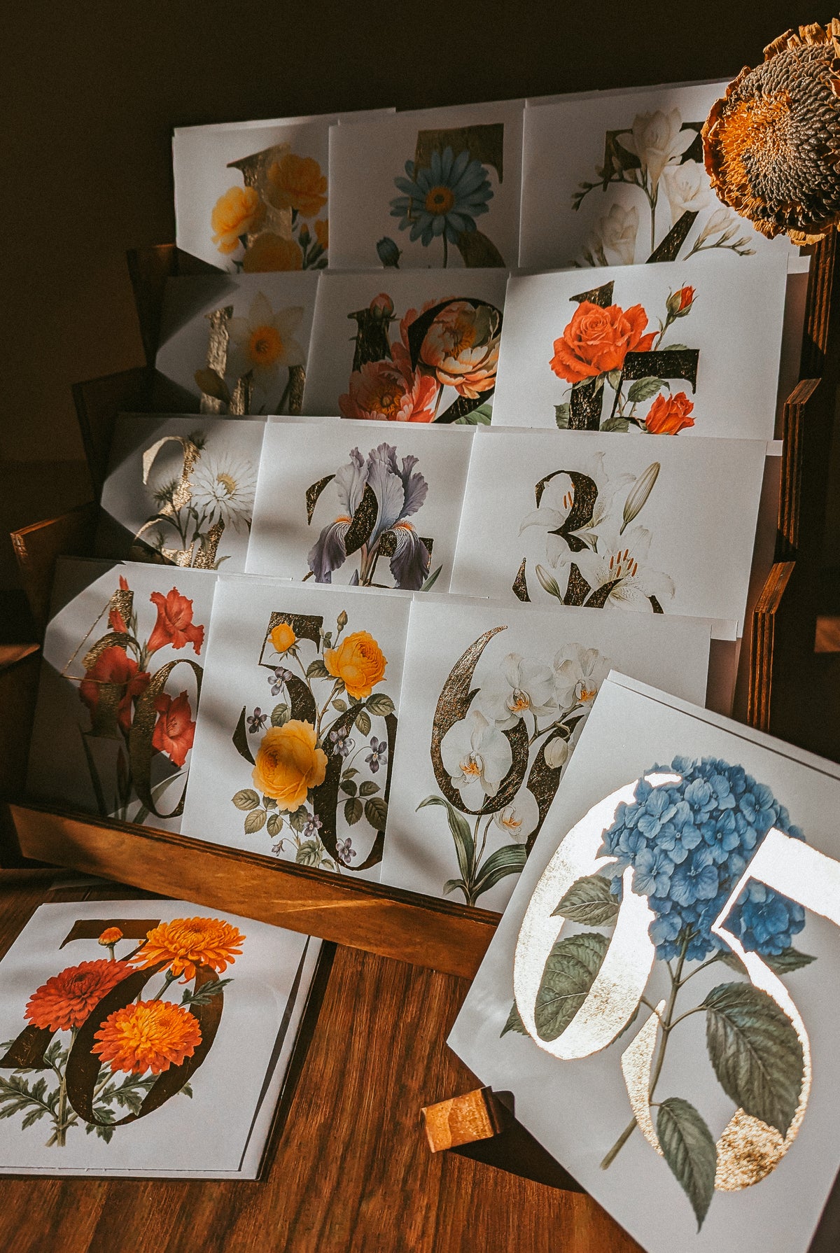 A styled flat-lay display of vintage-style botanical art cards featuring flowers and leaves in orange, blue, yellow, and white, arranged on a wooden surface with a woven textile in the corner.