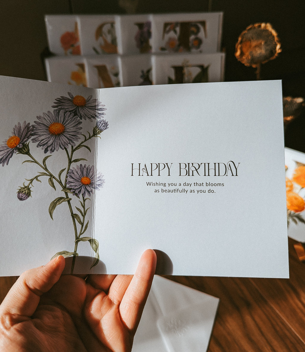 A hand holds an open greeting card with a floral illustration on the left panel and the words “HAPPY BIRTHDAY” on the right panel. The card is photographed in a warm indoor setting with blurred background decor and a wooden surface.
