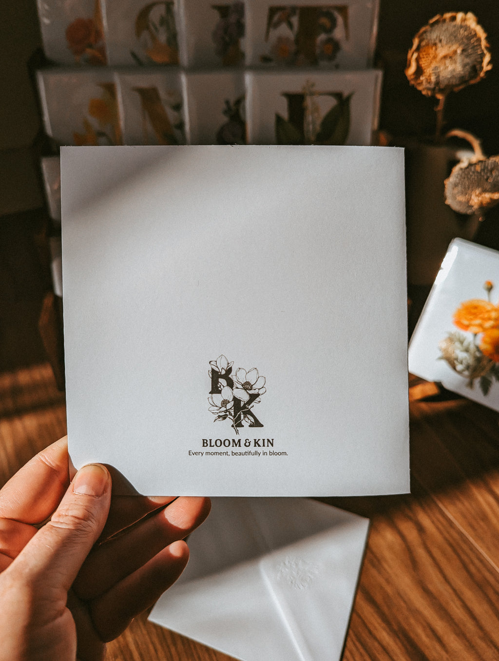 A hand holds a square white card featuring the black printed logo and text 'BLOOM & KIN' with a small floral illustration. The scene is set on a wooden surface with dried flowers and other branded cards or boxes in the background.