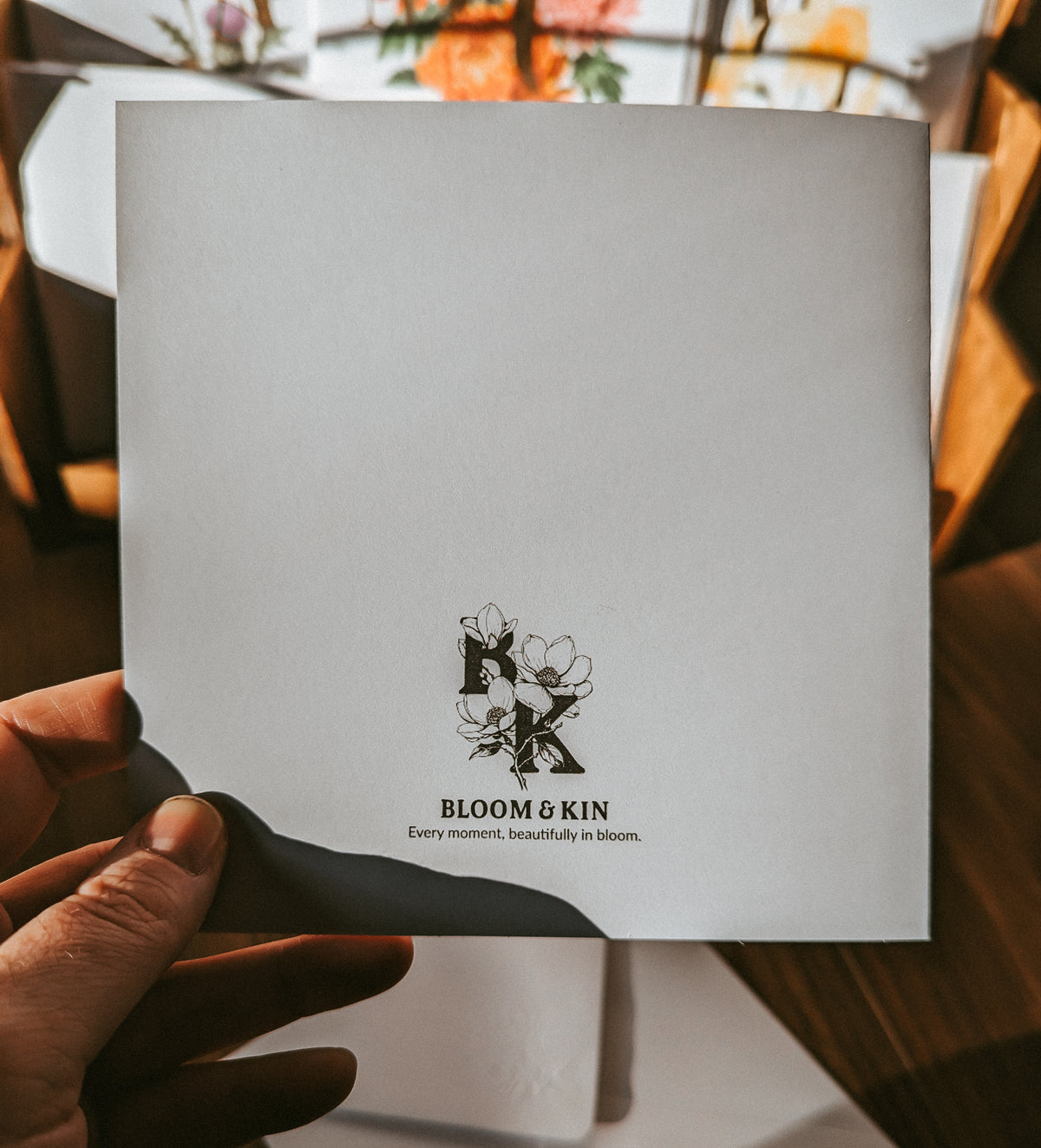 A hand holds a white square card or envelope with a black printed logo and the words 'BLOOM & KIN' and 'Every meaningful beautifully in bloom.' The background is a warm, softly blurred indoor setting with wooden surfaces and floral decor.