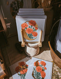 A stack of art cards featuring red carnation flowers on a gold initial 'C', displayed on small wooden stands in a styled indoor setting with dried plants and warm rustic decor.