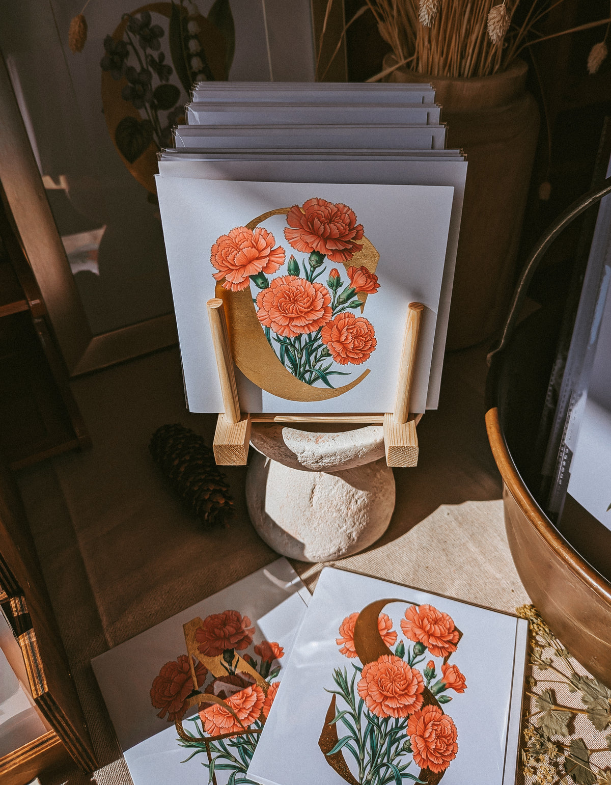 A stack of art cards featuring red carnation flowers on a gold initial 'C', displayed on small wooden stands in a styled indoor setting with dried plants and warm rustic decor.