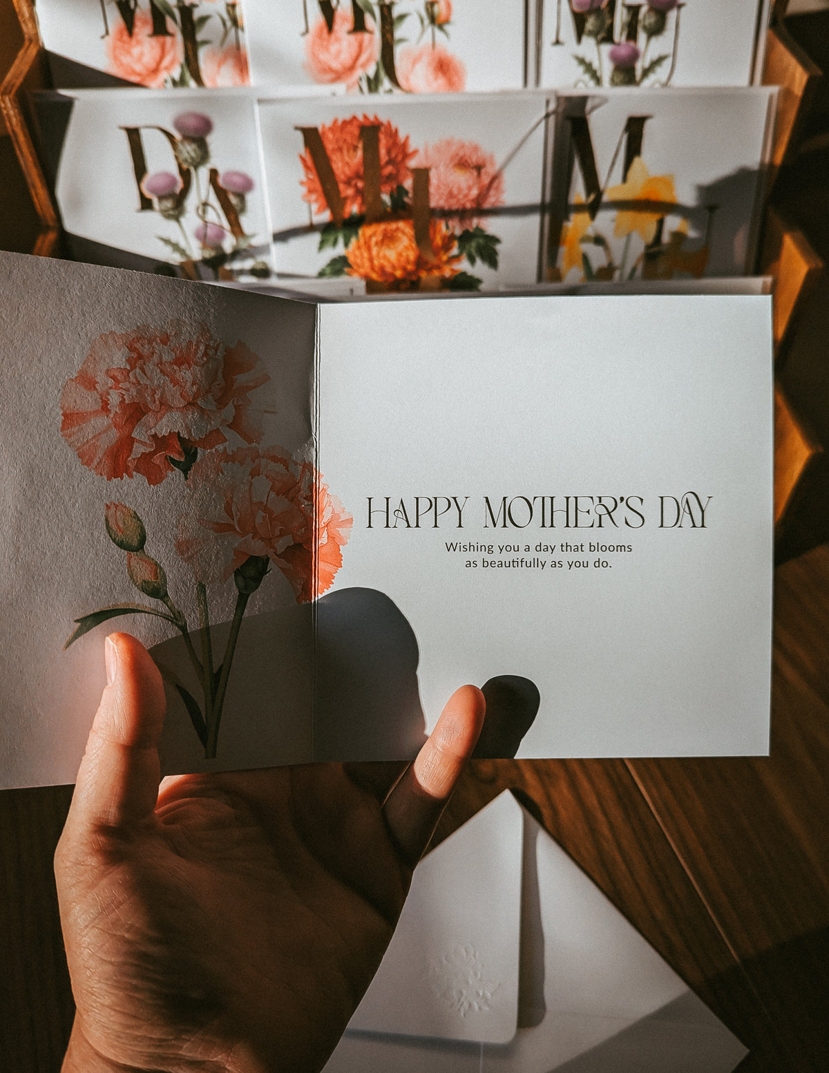 A hand holds an open Mother's Day greeting card with a watercolor floral design on the left panel and the text 'HAPPY MOTHER'S DAY' on the right. More cards are stacked in the background on a wooden surface.