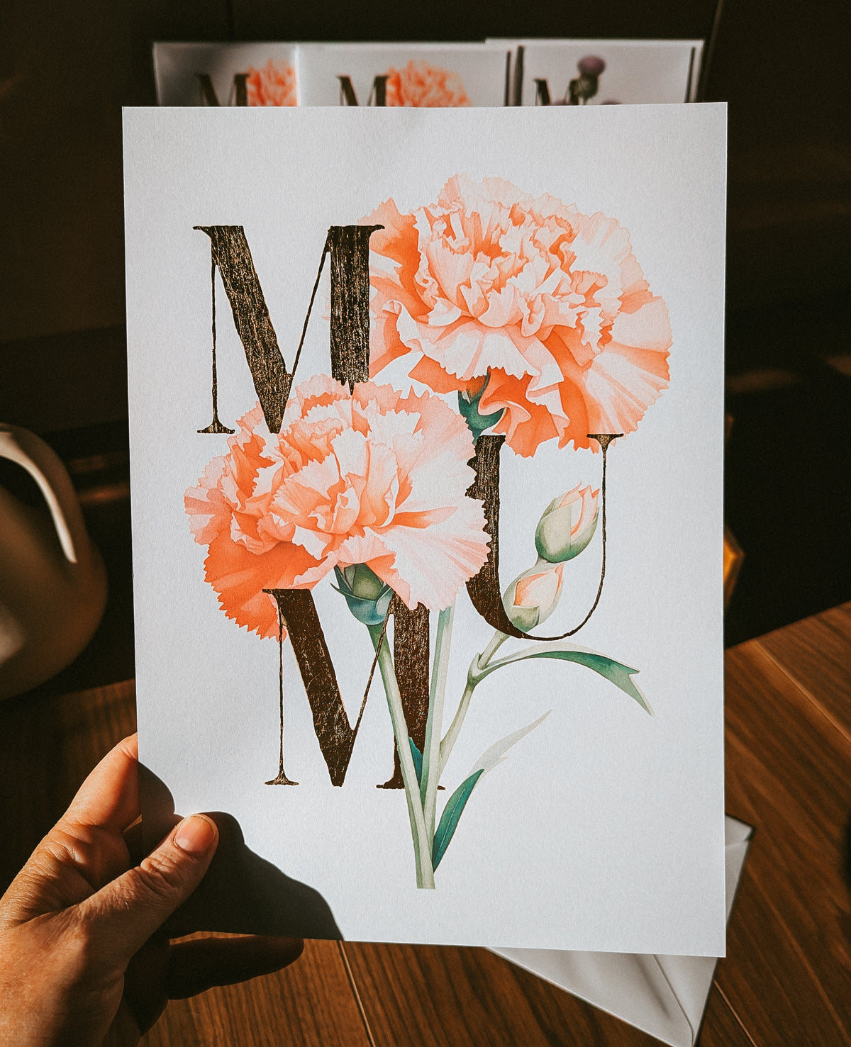 A hand holds a white print featuring a floral carnation design with the word 'MUM' in gold foil serif letters and two large peach-pink carnation flowers with green stems. The background is a warm, dark wood surface.