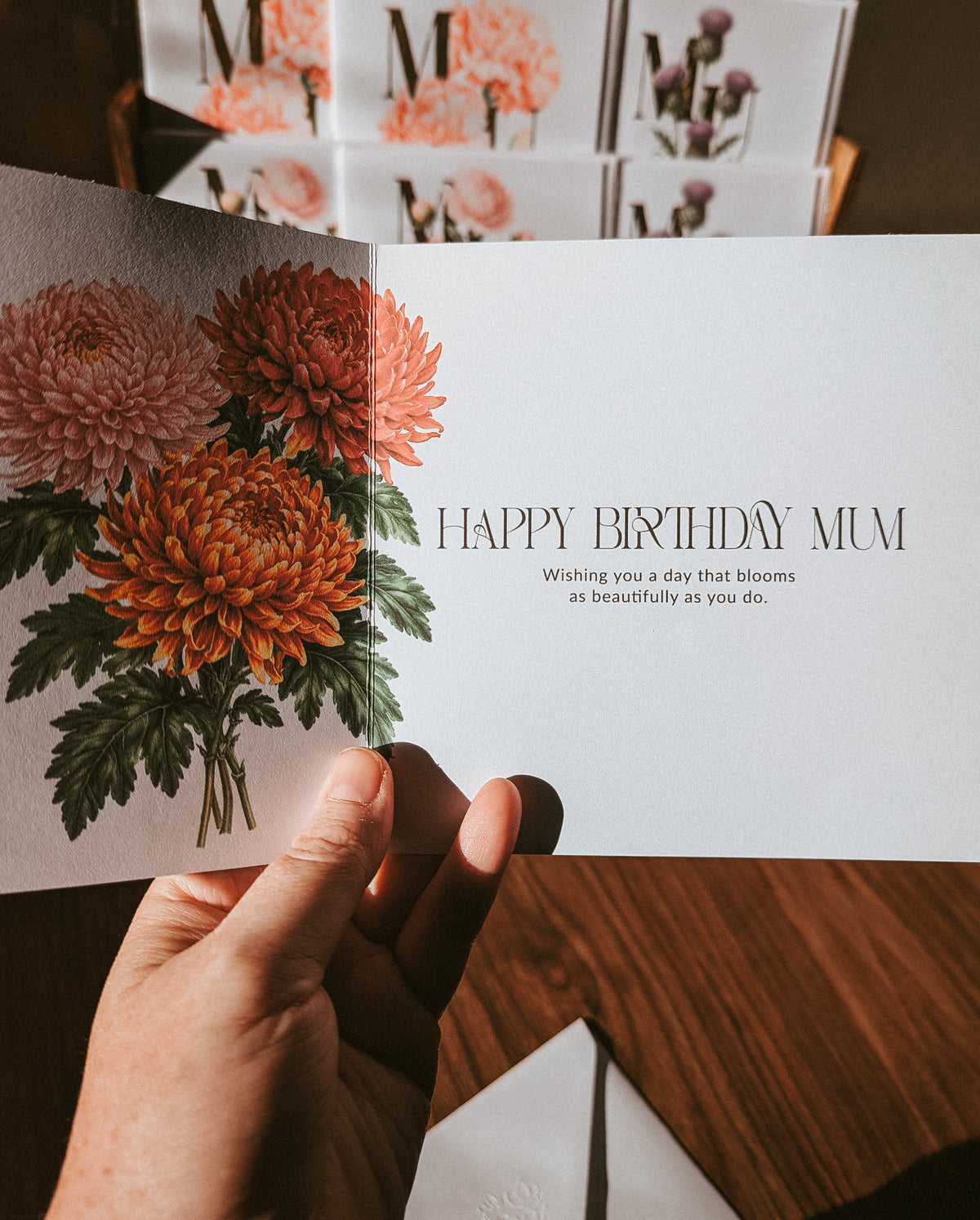 A hand holds an open floral birthday card with orange and pink chrysanthemums and the text 'HAPPY BIRTHDAY MUM' on a white card, with more matching cards stacked in the background on a wooden surface.