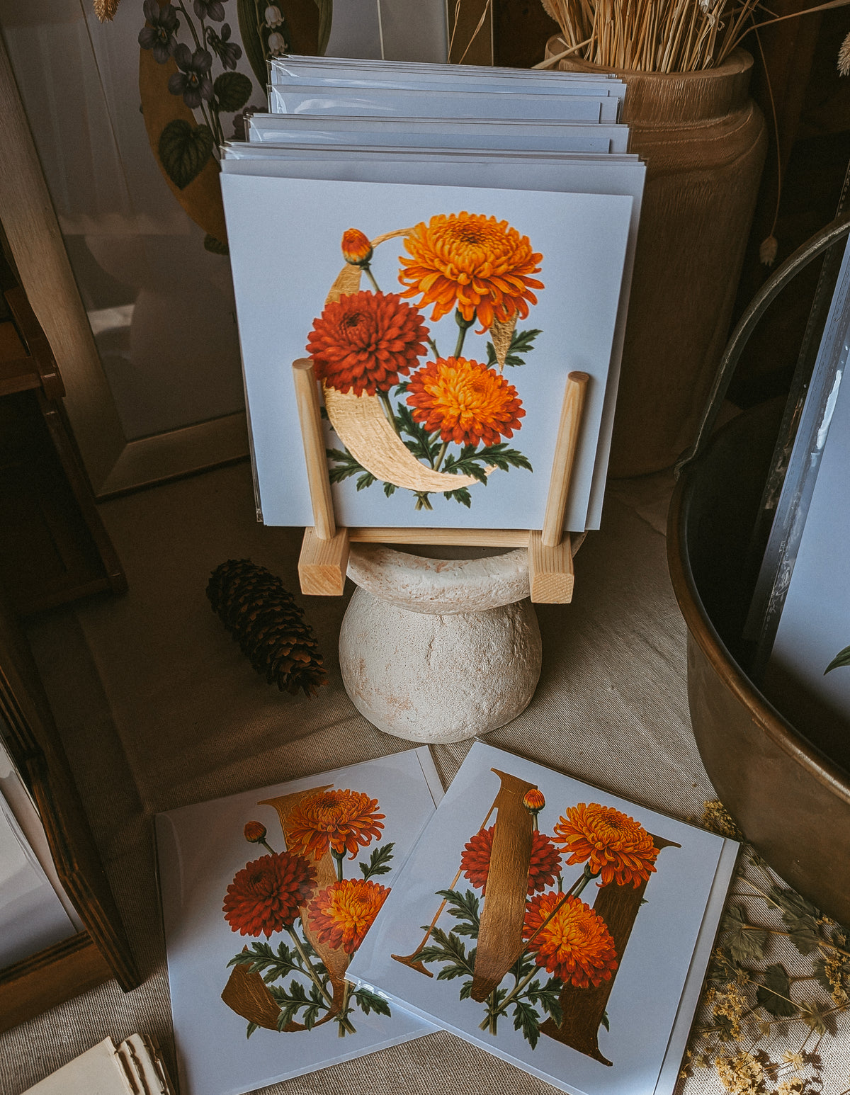 A stack of square art cards displayed on a small wooden stand, with two additional prints laid flat in front. The prints feature a vintage-style botanical illustration of orange and red chrysanthemum flowers with a large letter 'C'.