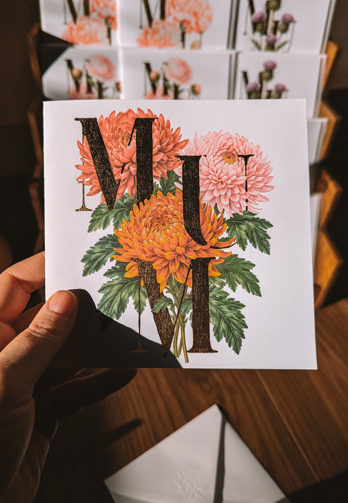 A hand holds a square white greeting card featuring a floral design with large gold foil letters spelling 'MUM'. The card has orange and pink chrysanthemums with green leaves.