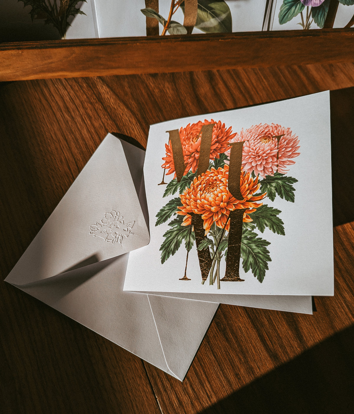 A square white greeting card with a vintage-style floral illustration featuring orange and pink flowers, green leaves, and large gold foil monogram letters, displayed on a wooden table next to a white envelope.