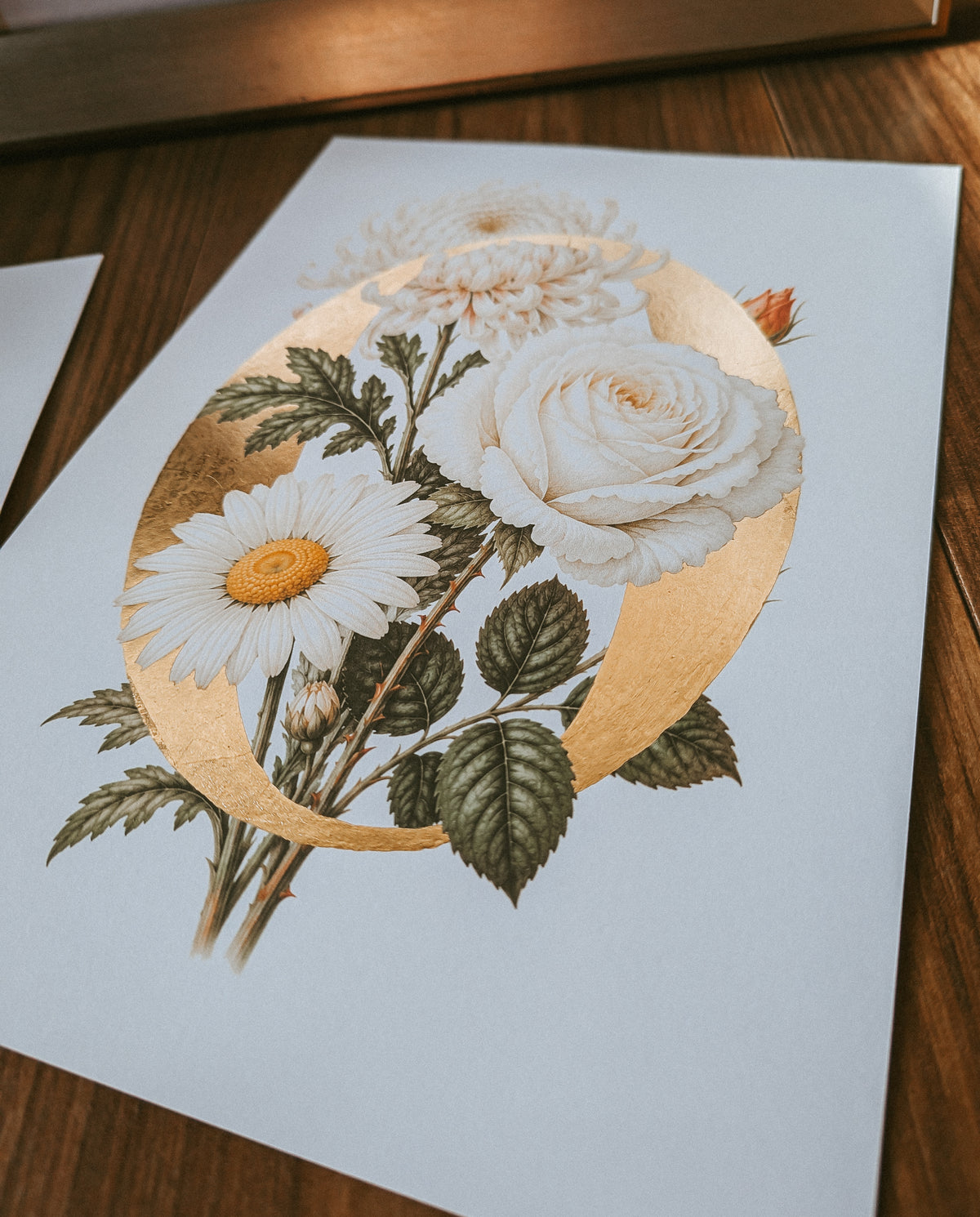 A botanical art print featuring white roses, daisies, and green leaves on a white sheet of paper with a large gold monogram, displayed on a wooden surface.