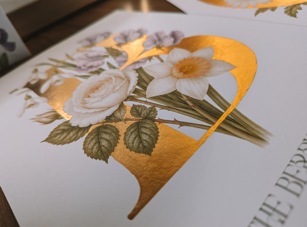 A close-up of a printed artwork featuring a large gold monogram letter B behind white and pale pink flowers with green leaves on a white background.