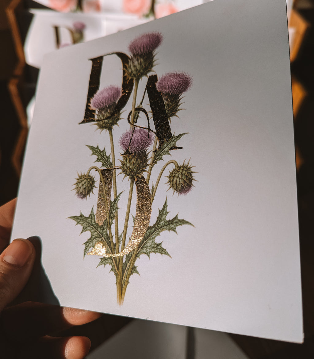 A hand holds a square white card featuring a botanical illustration of thistle flowers with the letters 'DAD' integrated into the design. The background is dark and softly blurred.