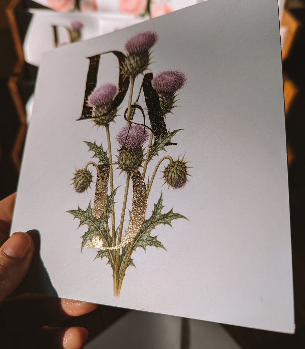A hand holds a square white card featuring a botanical illustration of thistle flowers with the letters 'DAD' integrated into the design. The background is dark and softly blurred.