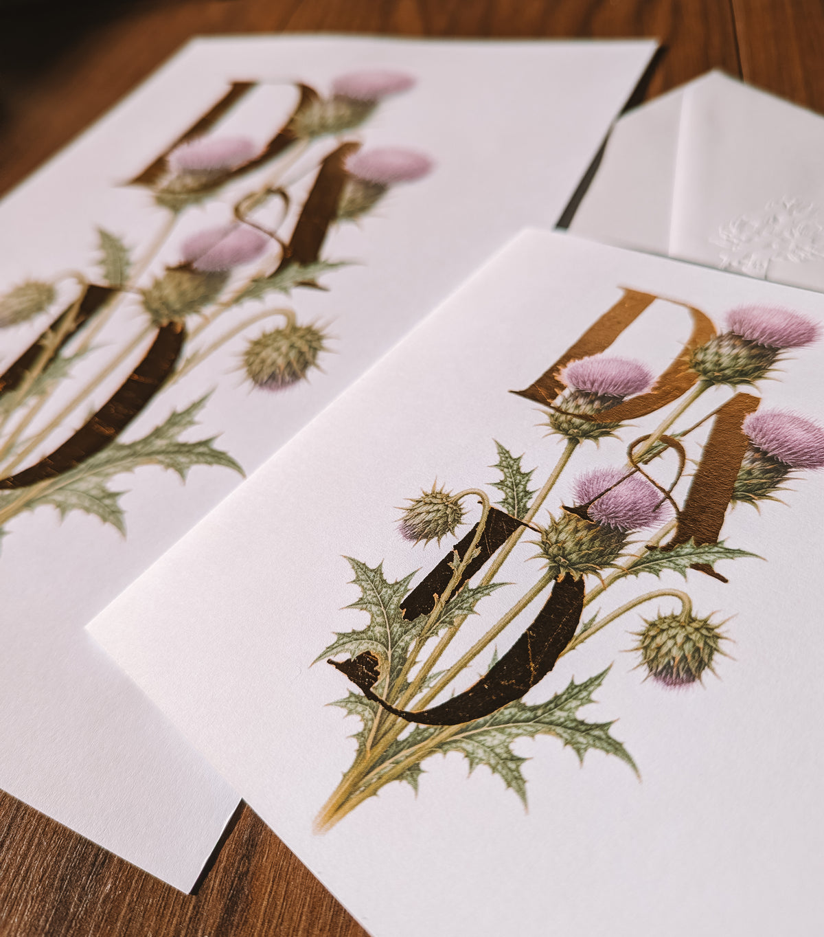 White stationery card & print featuring a vintage-style botanical monogram letterS 'Dad' decorated with thistle flowers, arranged on a dark wooden surface next to a white envelope.