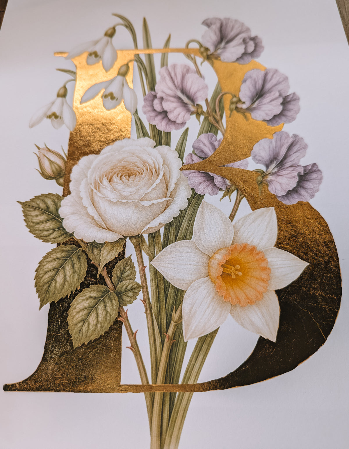 A decorative floral illustration featuring a large gold letter B adorned with white and purple flowers, green leaves, and a daffodil, set against a plain light background.